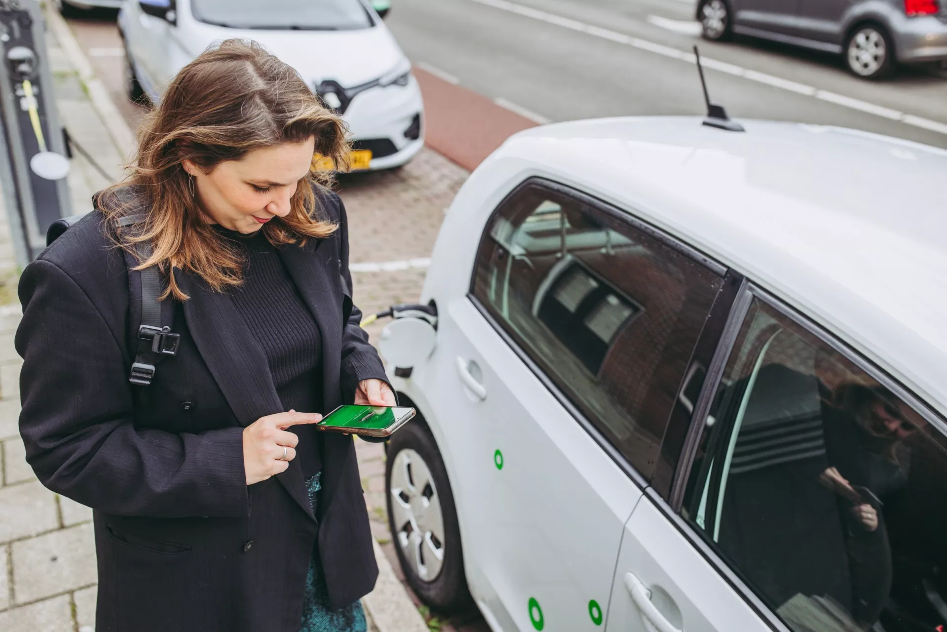 Vrouw gebruikt een smartphone naast een geparkeerde elektrische auto.